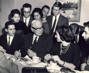 Bernard Baruch surrounded by a group of college students. Baruch is seated at a table with two students by his side. There are teacups on a table in front of them.